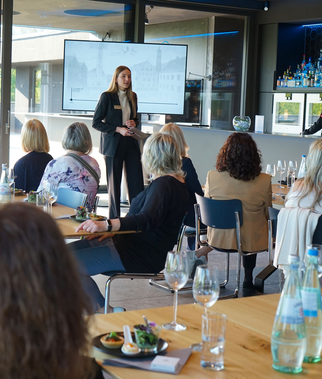 Frau in schwarzem Anzug steht vor einer Gruppe sitzender Frauen in einem modernen Raum mit Tischen, Gläsern und Flaschen                               
