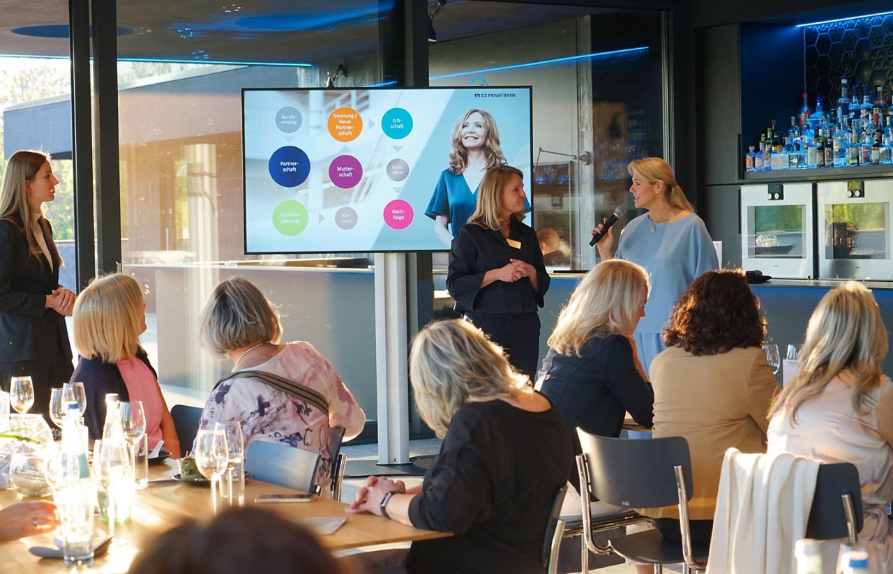 Mehrere Frauen sitzen an Tischen und hören zwei Frauen zu, von denen eine mit Mikrofon vor einem Bildschirm mit bunten Kreisen spricht                               