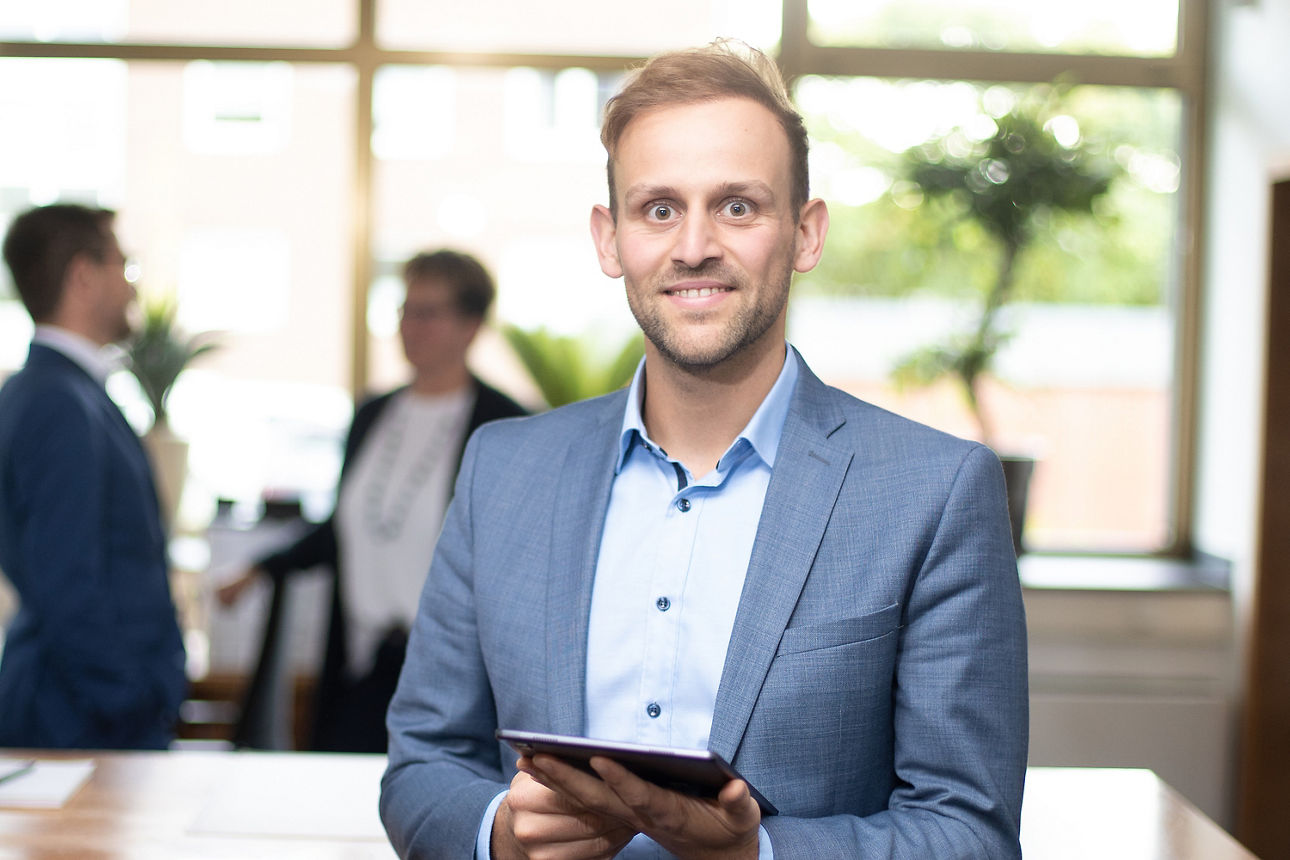 Mitarbeiter Tom Weber mit einem Tablet in der Hand vor dem Schalter in unseren Räumlichkeiten in Gahlen - im Hintergrund sind zwei weitere Mitarbeiter verschwommen im Bild