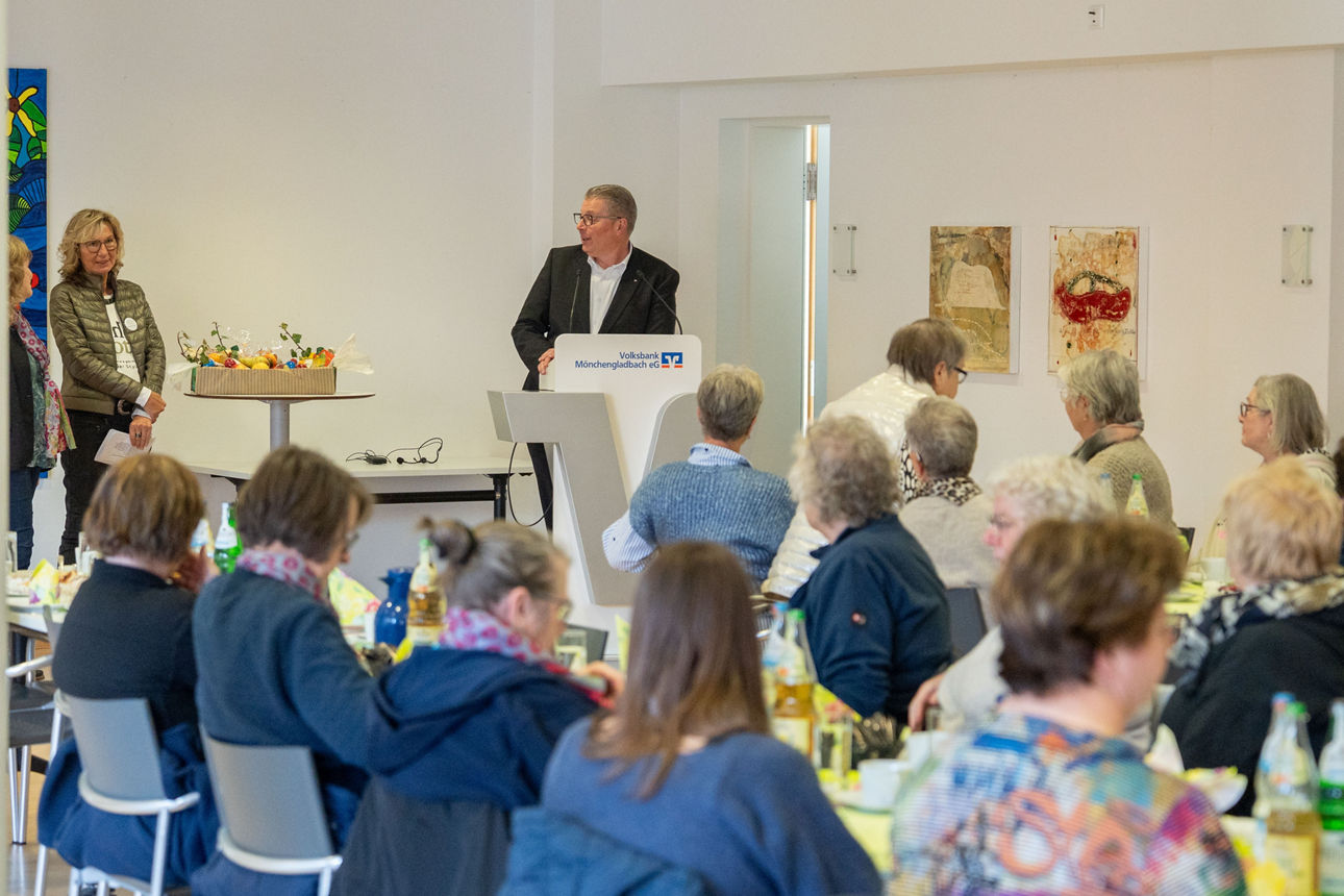 Das Foto zeigt eine Gruppe von Menschen, die in einem Bankgebäude sitzen. Vorne stehen mehrere Leute am Sprecherpult.