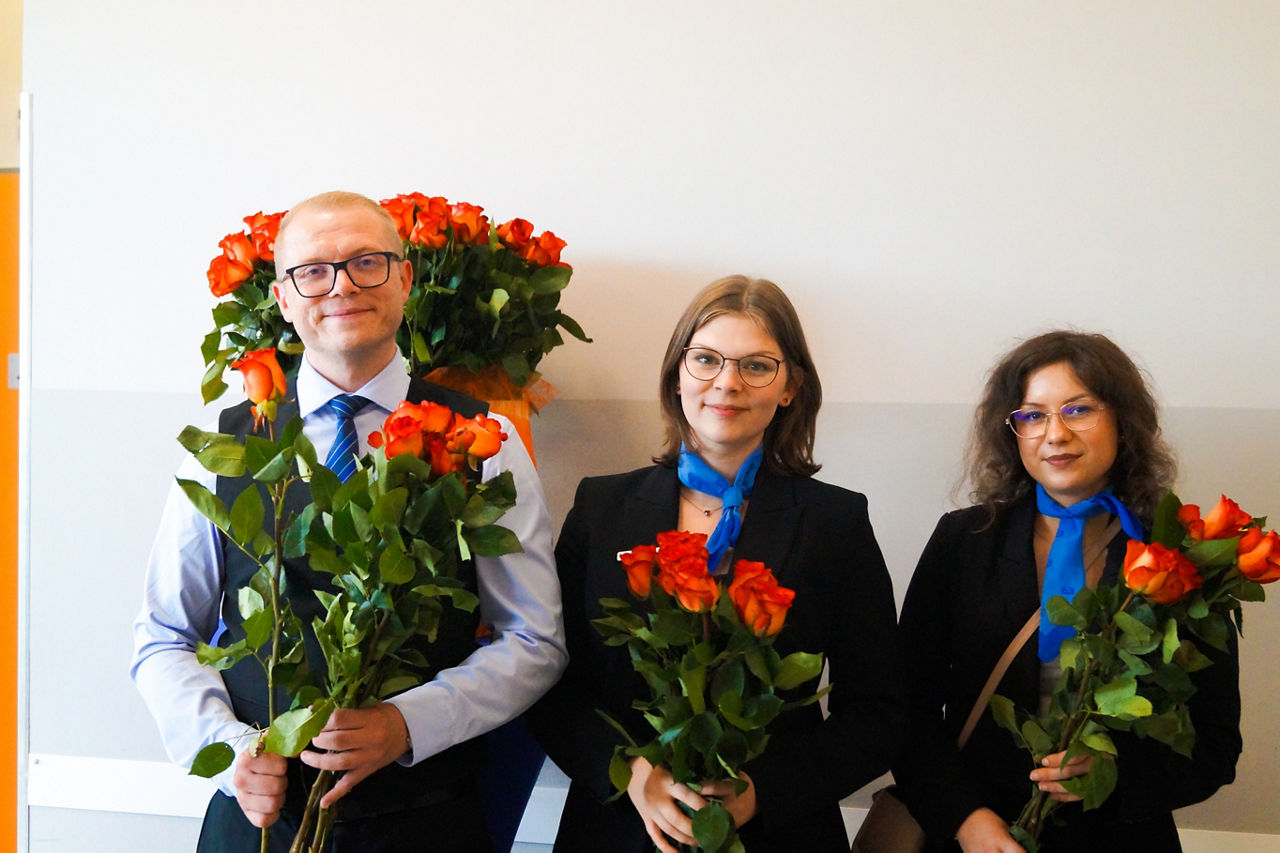 Zwei Damen und ein Herr mit Rosen vor einer weißen Wand. 