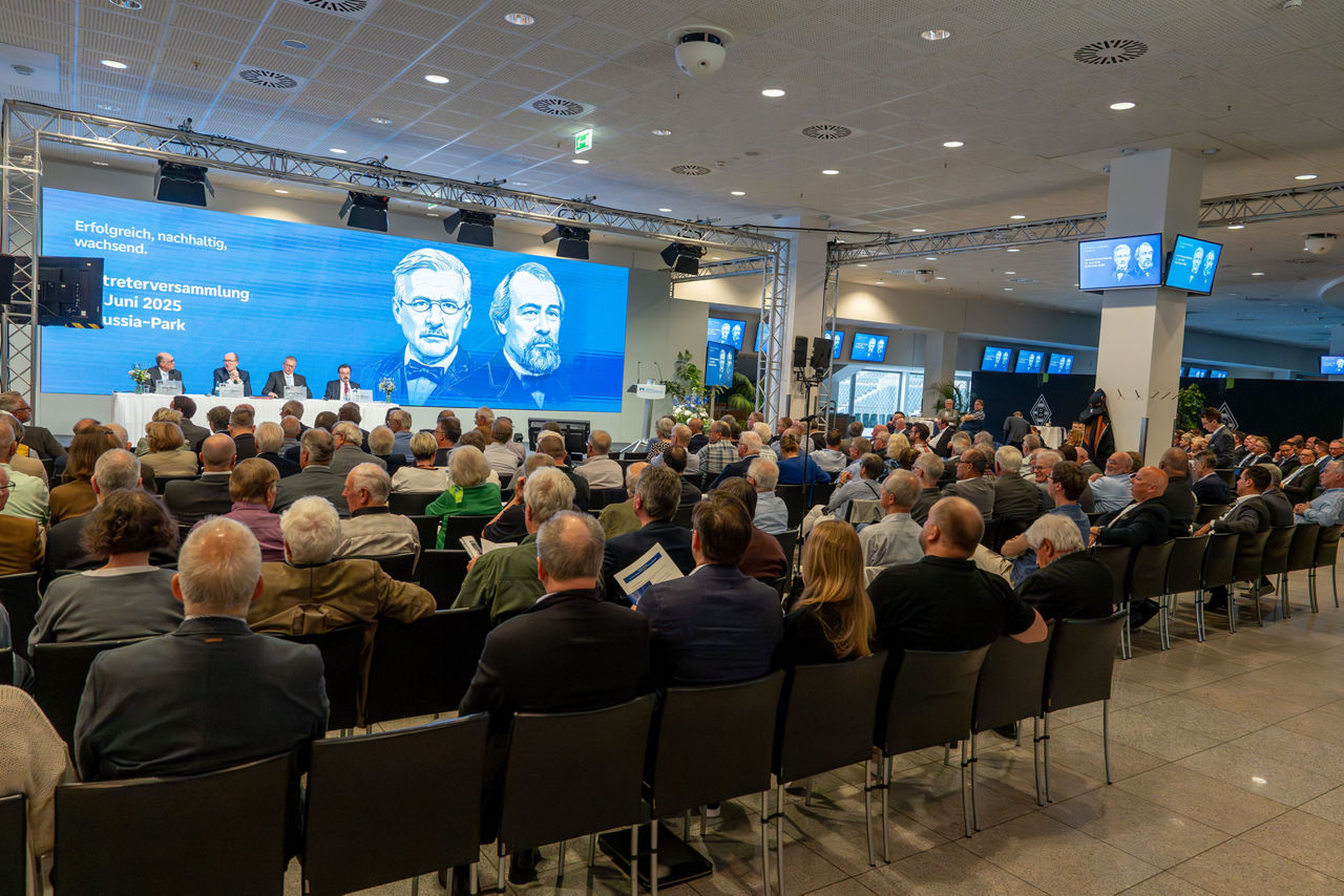Eine größere Versammlung in einem Konferenzraum. Viele Menschen sitzen in Stuhlreihen und blicken nach vorn zur Bühne