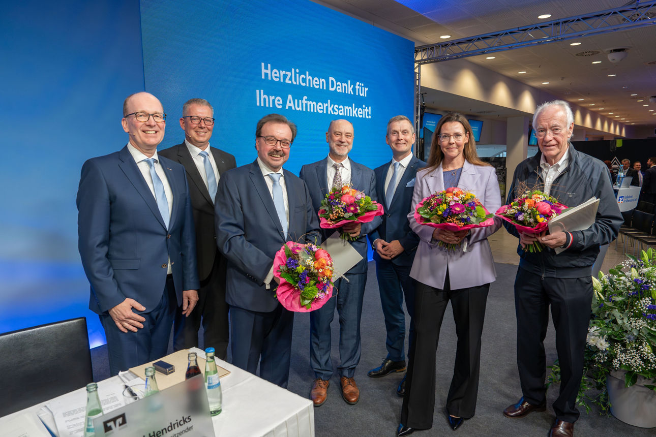 Auf dem Foto stehen sieben Personen in einer Reihe auf einer Bühne oder Präsentationsfläche. 