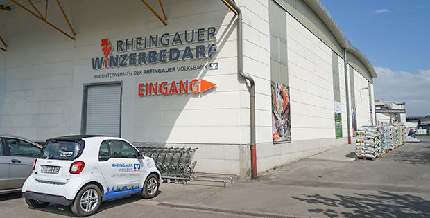 Ein eSmart der Rheingauer Volksbank steht vor einer Lagerhalle, auf der das Logo des Rheingauer Winzerbedarfs zu sehen ist.