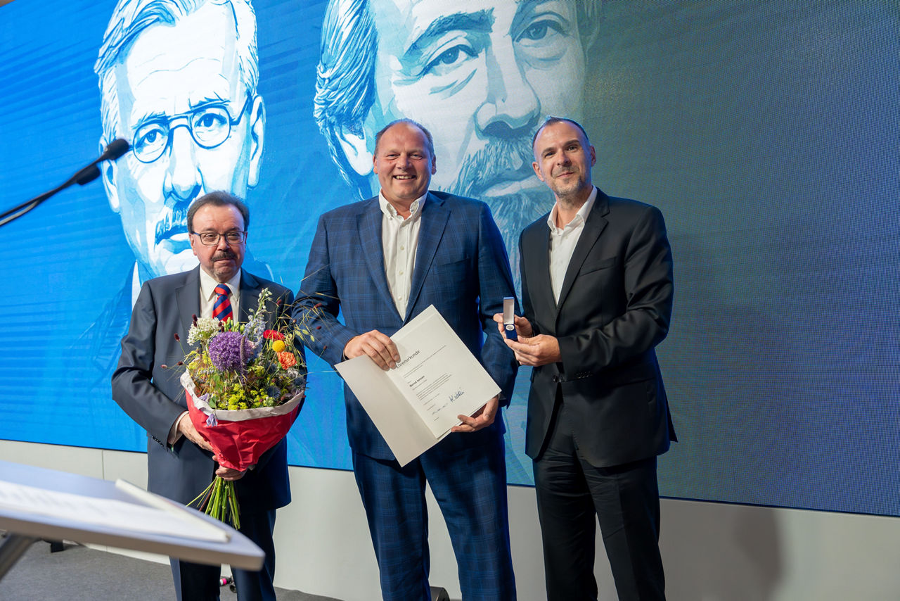 Drei Männer stehen auf einer Bühne vor einem großen Bildschirm mit zwei gezeichneten Porträts. 