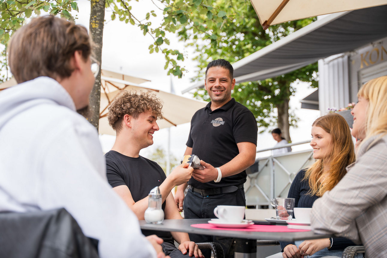 Eine Gruppe sitzt draußen am Tisch im Cafe und ein Mann bezahlt mit seiner ec Karte beim Kellner