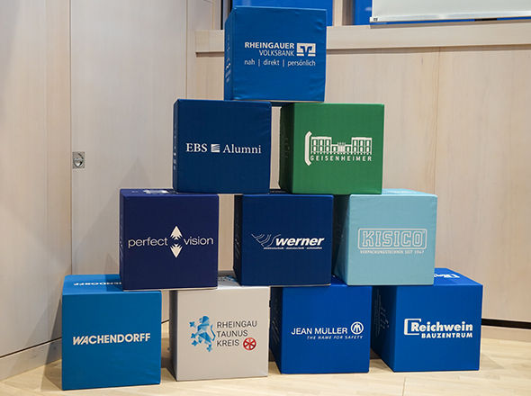 Das Bild zeigt einen Stapel aus zehn bunten Würfeln mit Firmenlogos und Namen. Die Würfel sind in Blau-, Grün- und Beigetönen