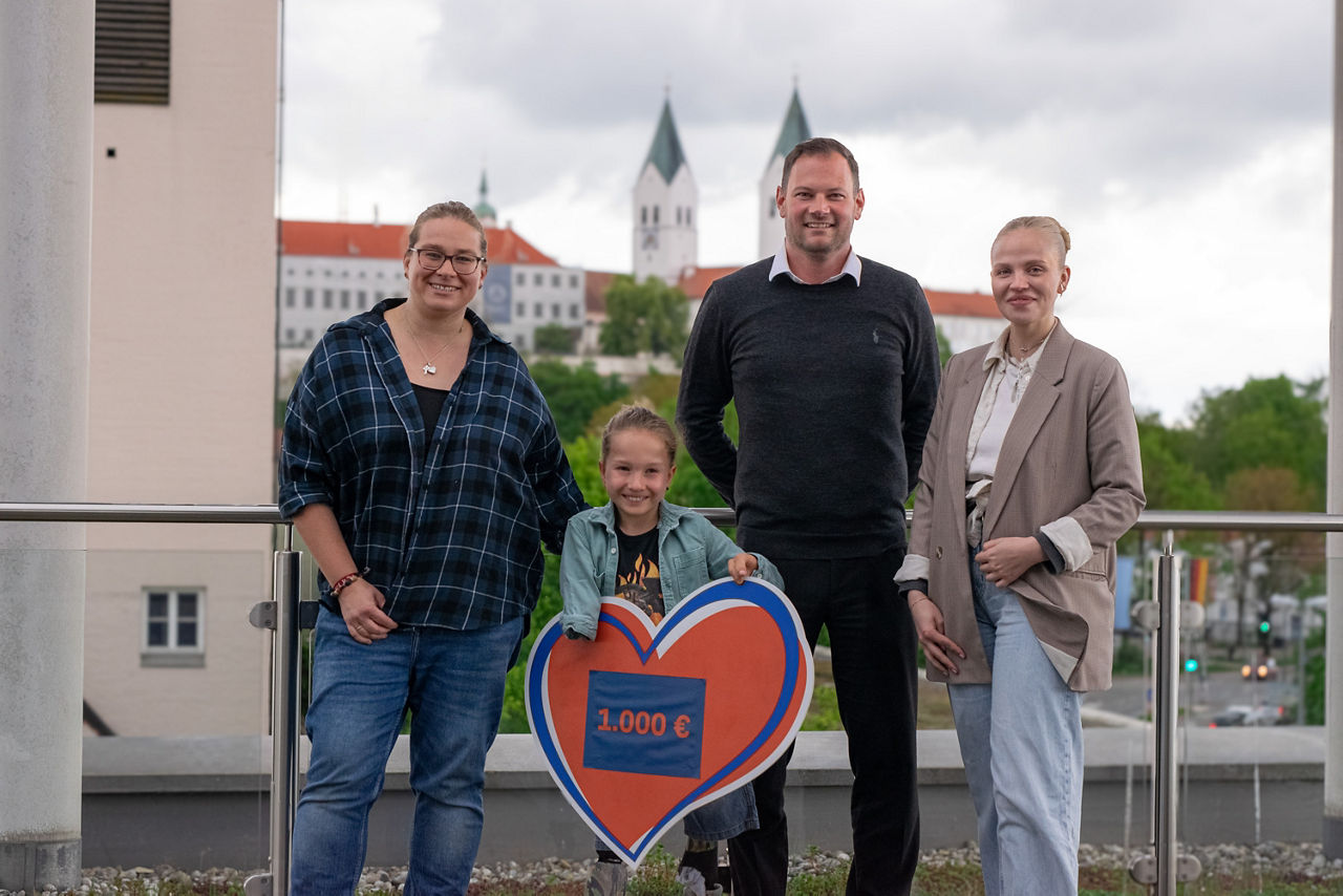 Vier Personen stehen auf Dachterrasse der Freisinger Bank mit Dom im Hintergrund; zwei Frauen und ein Mann umrahmen kleinen Bub der ein Spendenherz mit Aufschrift 1.000 Euro hält