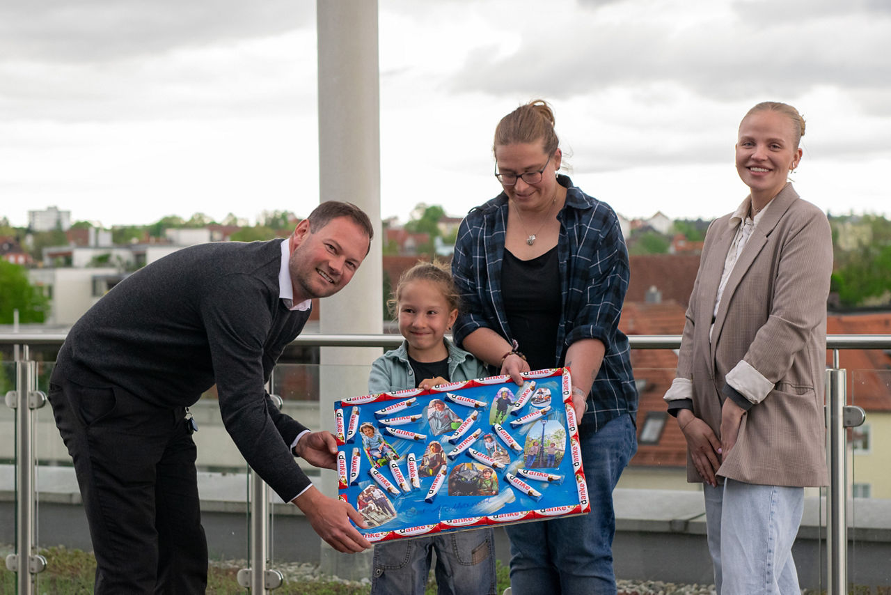 Gruppe von drei Erwachsenen und einem Jungen stehen auf Dachterrasse; der Bub, seine Mutter und ein Mann halten eine selbstgebastelte Kollage in die Kamera