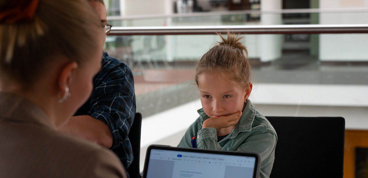 Kleiner Junge stützt sein Kinn auf eine Hand und schaut zu seinem Gegenüber, einer Frau von hinten fotografiert mit Laptop