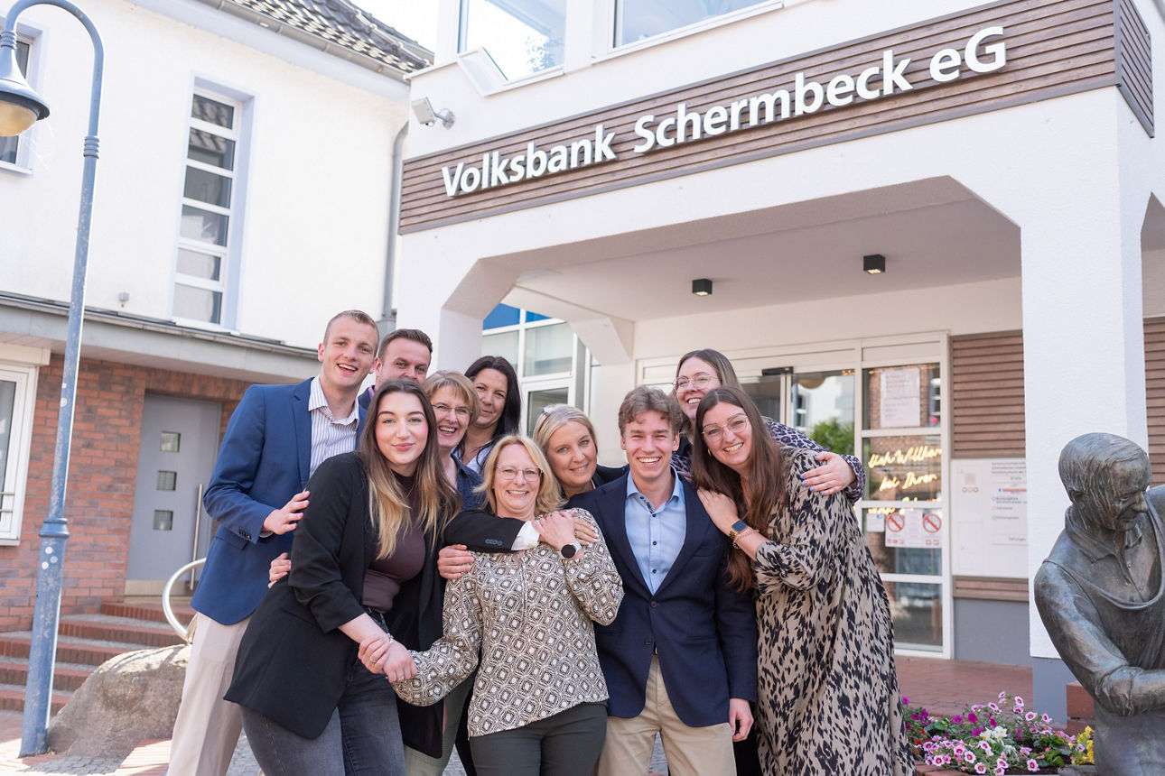 Mitarbeitende der Volksbank Schermbeck stehen lachend Arm in Arm vor der Bank.