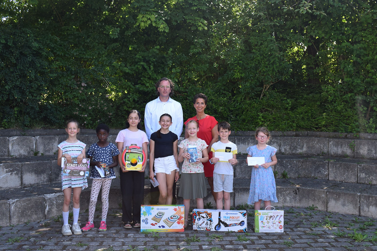 Zwei Erwachsene Personen stehen hinter Kindern auf einem Schulhof. Die Kinder halten Spiele in der Hand
