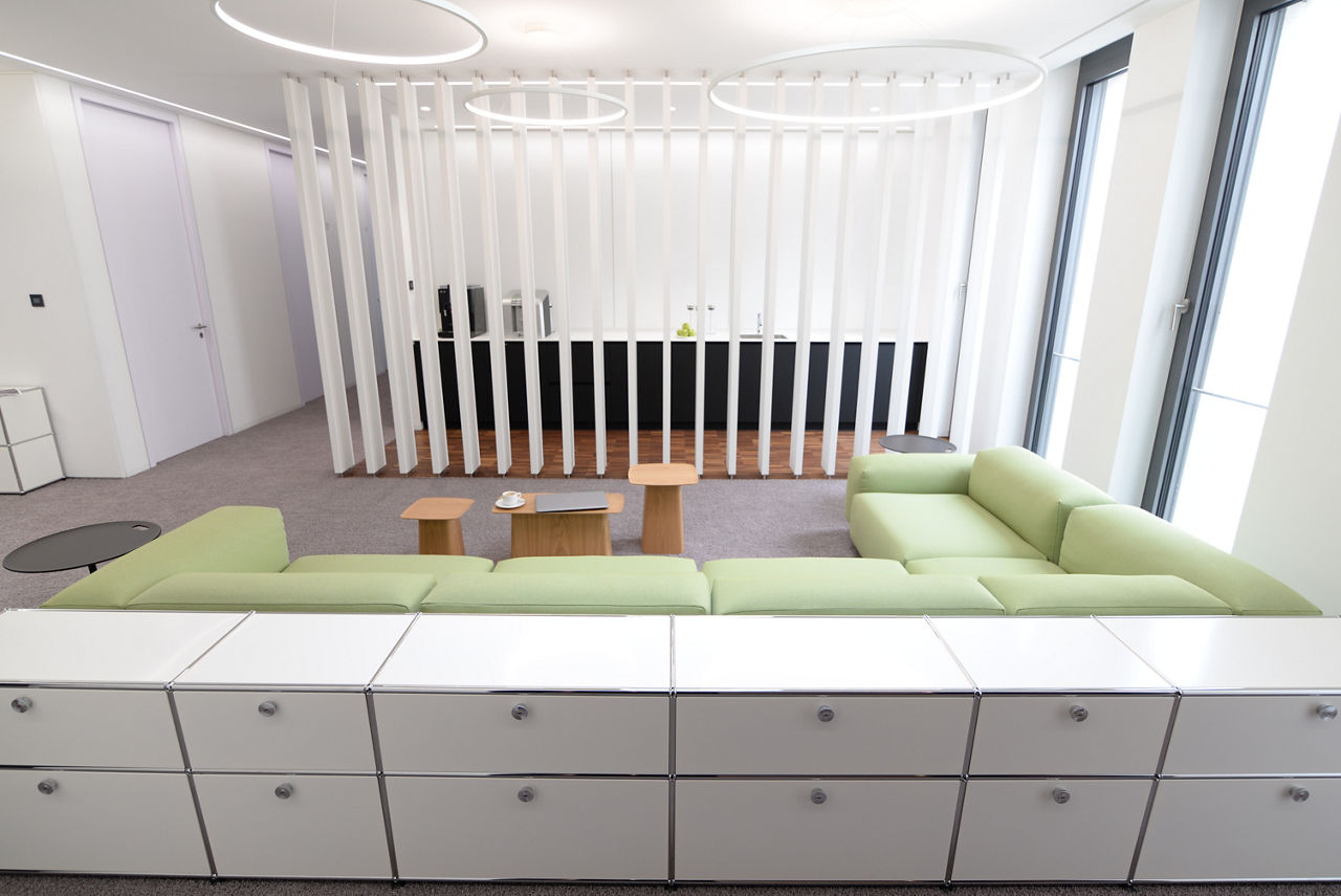 Eine Reihe weißer Sideboards vor eine hellgrünen Couch, der Blick geht auf eine transparente Wand, hinter der eine Küchenzeile zu sehen ist