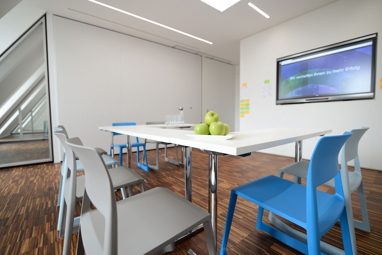 Raum mit bunten Stühlen, einem Besprechungstisch und einem großen Monitor an der Wand.