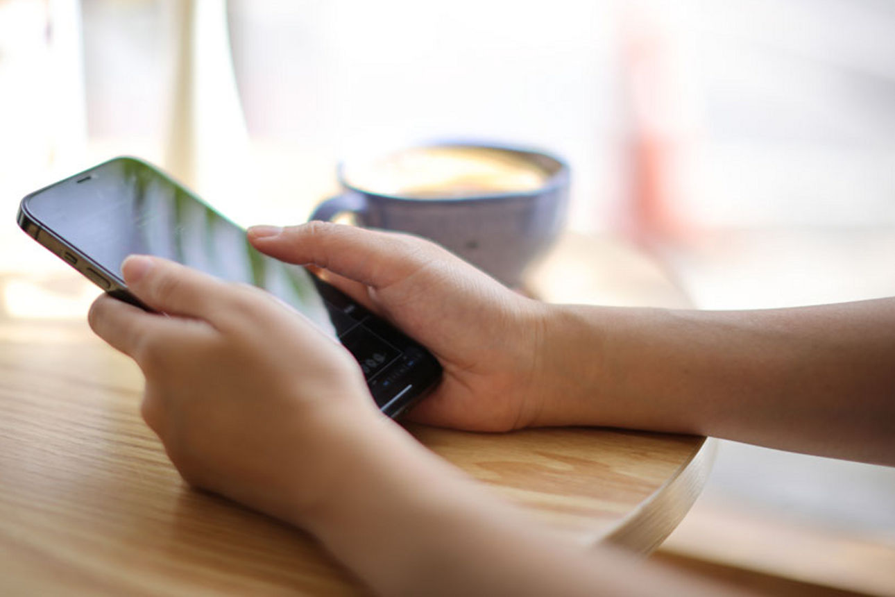 Zwei Hände halten ein Handy  in der Hand, nebendran steht eine Kaffeetasse