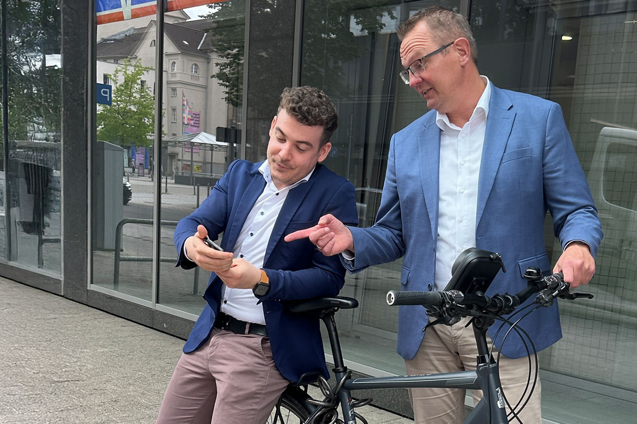 M. Ascione und G. Danzebrink lehnen an einem Fahrrad und schauen zusammen auf ein Handy