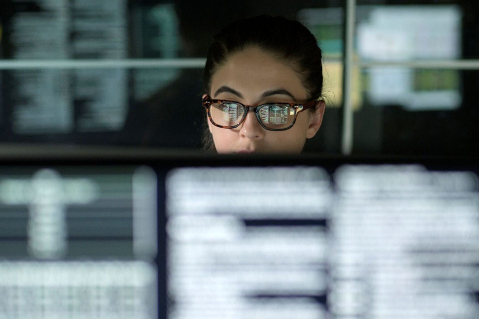 Eine Frau mit Brille sitzt vor einer Reihe mit Bildschirmen