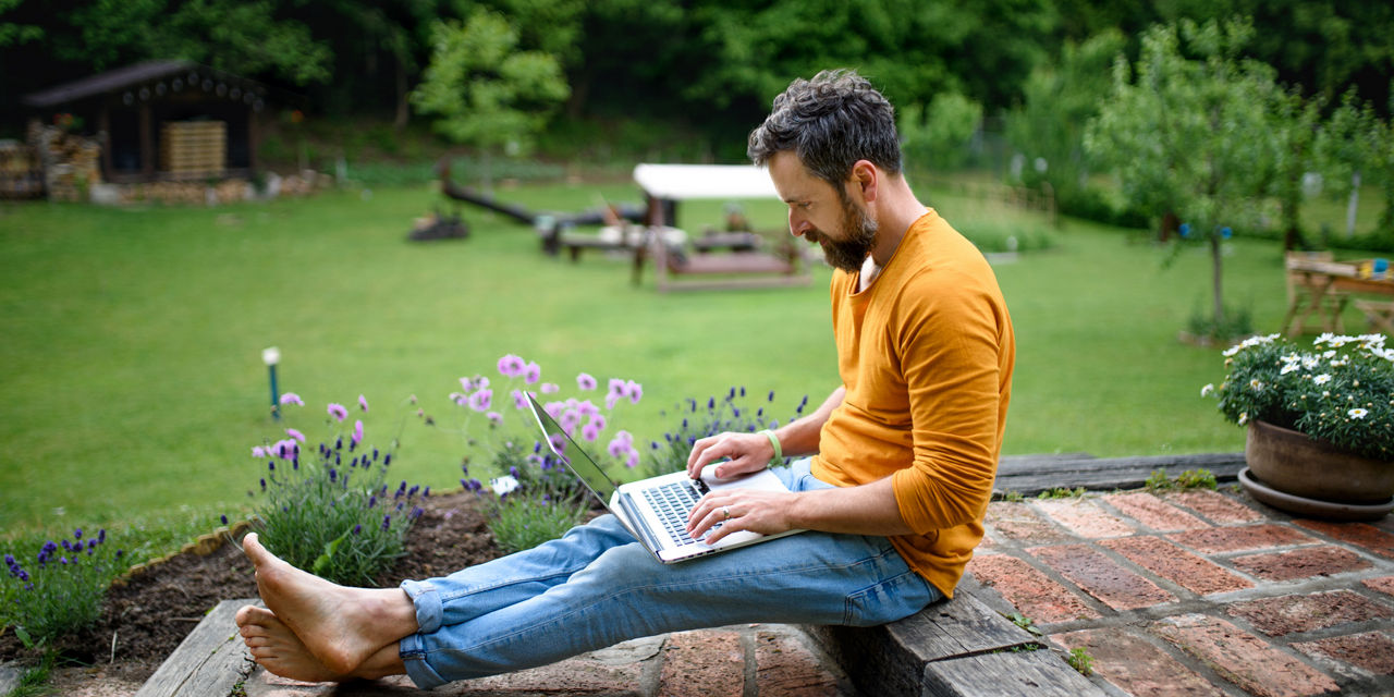 Person sitzt im Garten, arbeitet am Laptop, trägt ein orangefarbenes Shirt und blaue Jeans.