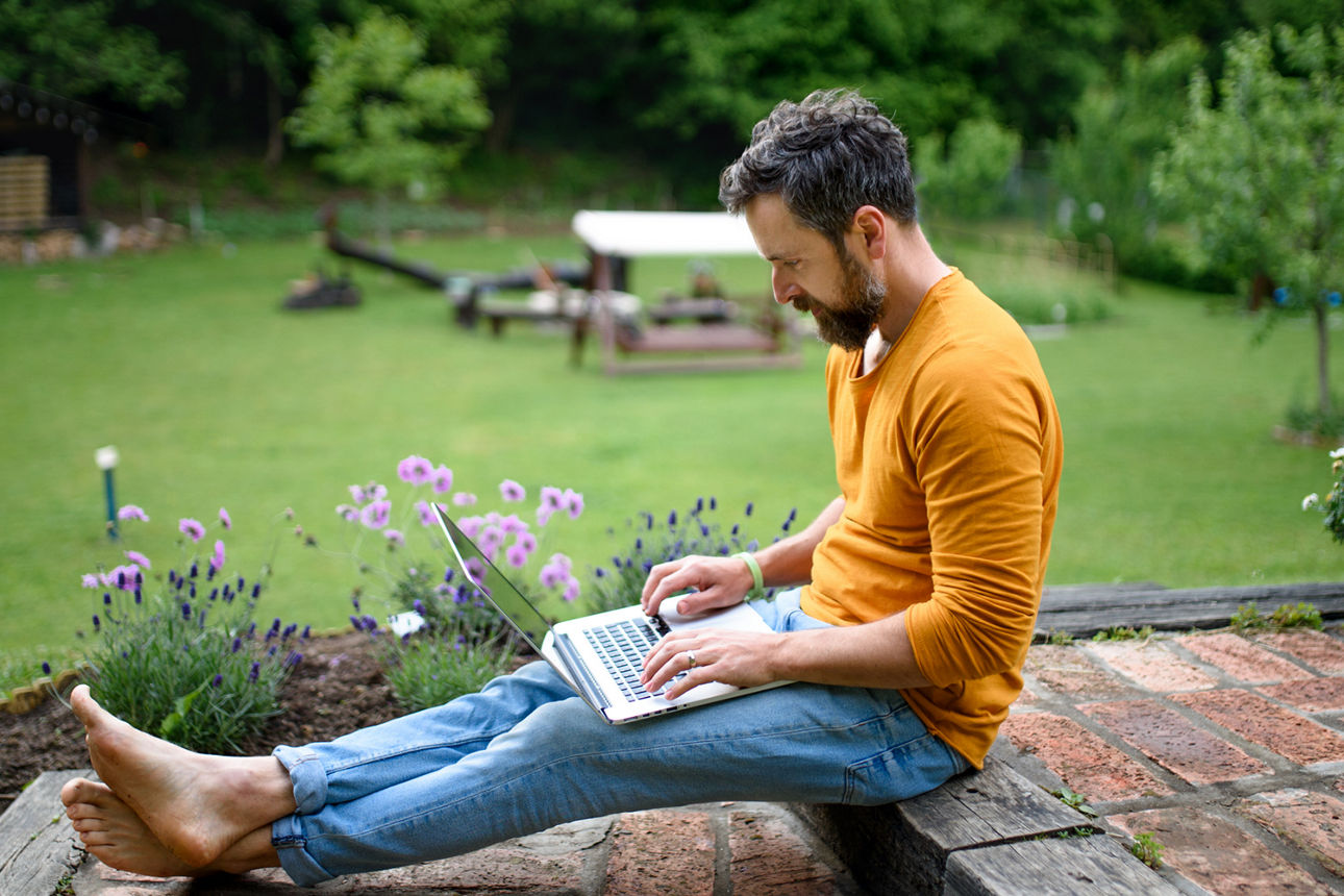 Person sitzt im Garten, arbeitet am Laptop, trägt ein orangefarbenes Shirt und blaue Jeans.