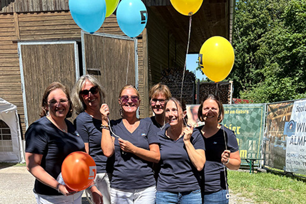 Frauen stehen in der Sonne, halten Luftballons in den Händen und lachen.