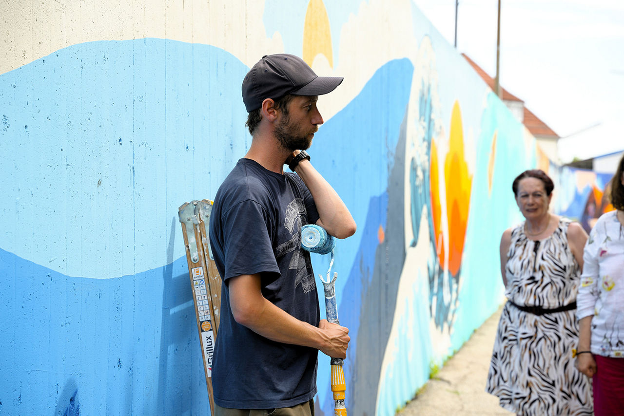 Junger Mann mit kurzen dunklen Haaren und Bart in dunkelblauem T-Shirt steht vor frisch bemalter Mauer mit Farbrolle in der Hand