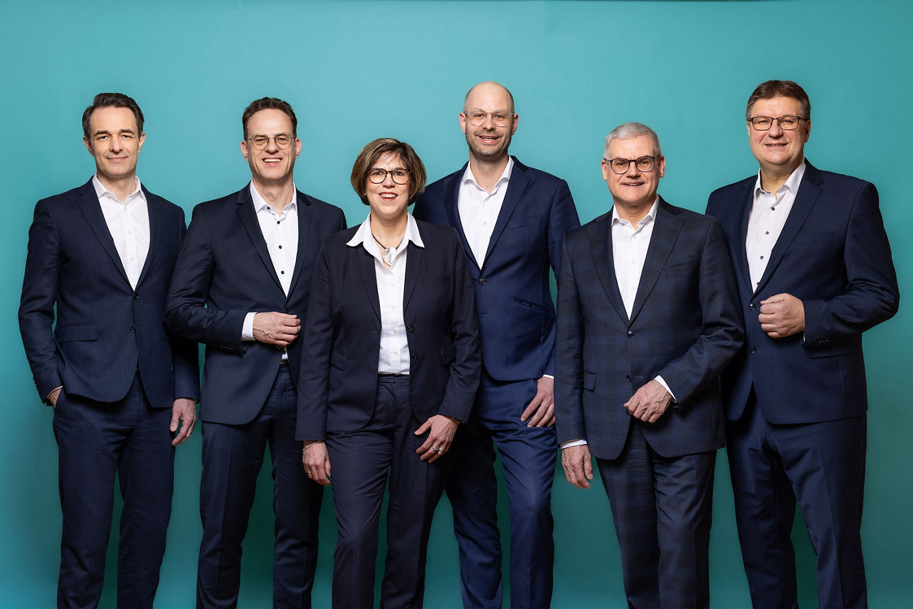 Der fünfköpfige Vorstand der neu entstehenden VR RegionalBank nebeneinanderstehend. Der Hintergrund ist petrolfarben.