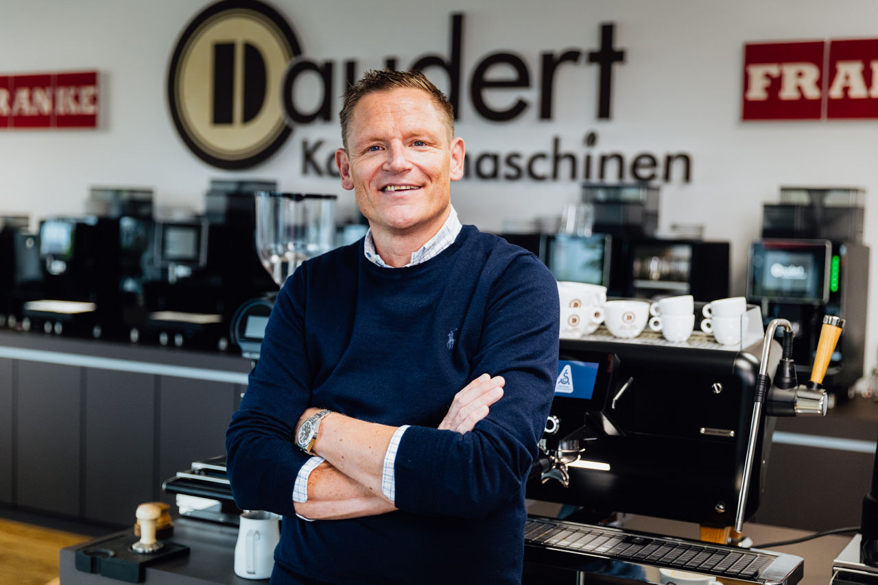 Innenaufnahme Daudert Kaffeemaschinen mit mehreren professionellen Kaffeemaschinen und Kaffeemühlen, im Vordergrund steht René Daudert vor einer Espressomaschine.