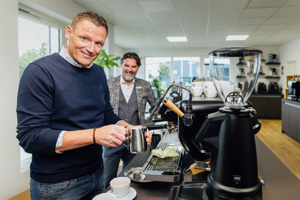 René Daudert und Berater Oliver Brust  stehen im Showroom vor einer Kaffeemaschine und halten Kaffeetassen, im Hintergrund sind Maschinen und Arbeitsflächen sichtbar