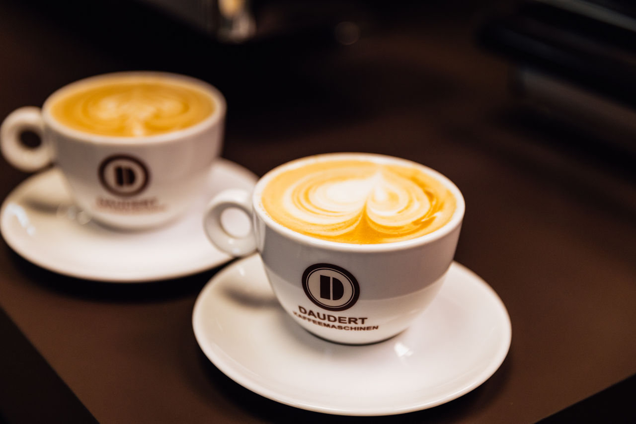 Zwei gefüllte Kaffeetassen mit Milchschaum auf Untertassen, abgestellt auf einer dunklen Oberfläche.