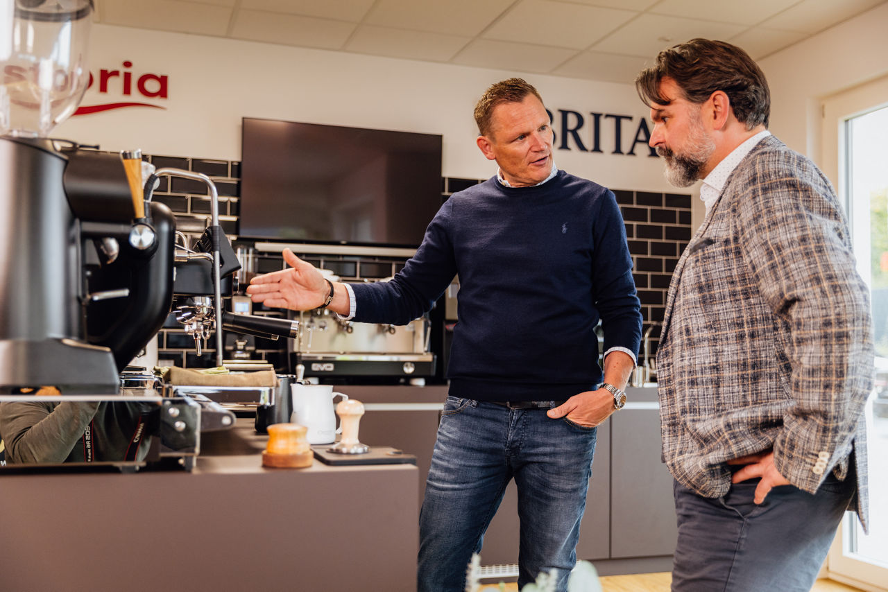 René Daudert und Berater Oliver Brust stehen neben einer Espressomaschine, eine Person zeigt auf ein Gerät während des Austauschs.