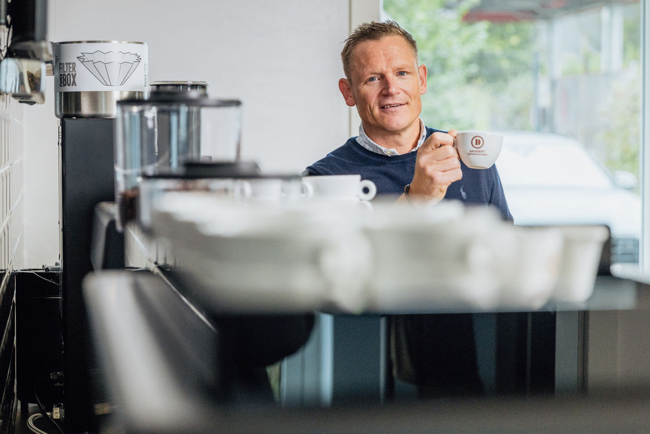 René Daudert hält eine gefüllte Kaffeetasse, im Vordergrund ist ein Regal mit Tassen und Kaffeemaschinen unscharf zu erkennen.