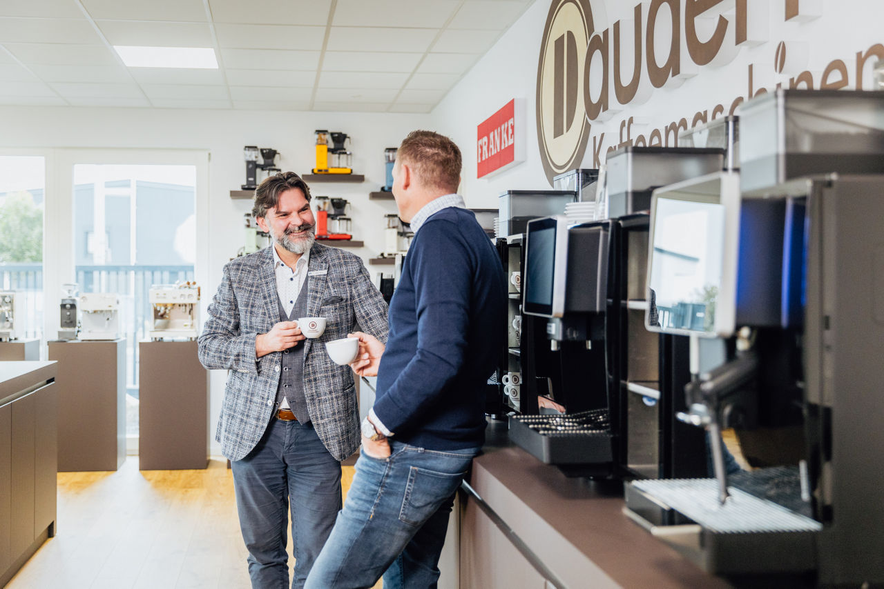 René Daudert und Berater Oliver Brust im Gespräch vor einer Reihe professioneller Kaffeevollautomaten in einem hellen Ausstellungsraum.