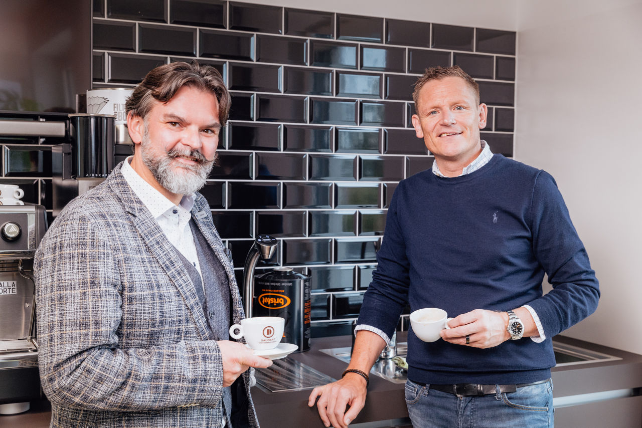 Berater Oliver Brust und René Daudert stehen vor einer Kaffeemaschine und halten Kaffeetassen, im Hintergrund sind Maschinen und Arbeitsflächen sichtbar.