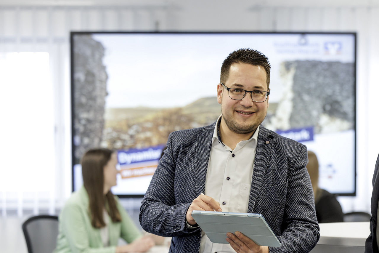 Bankberater steht vor einem großem Bildschirm und hält ein blaues Tablet in der Hand.