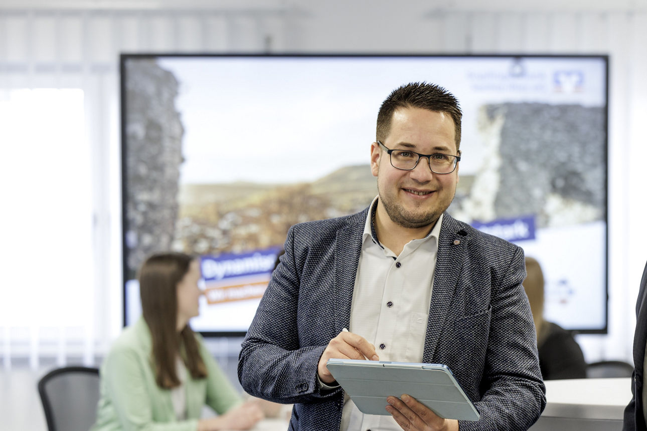Bankberater steht vor einem großem Bildschirm und hält ein blaues Tablet in der Hand.