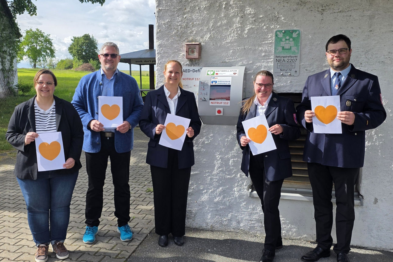 Fünf Personen halten orangefarbene Herzen in den Händen. Sie stehen vor einem Defibrillator an einer Hauswand.