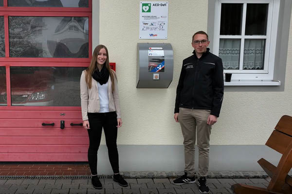 Zwei Bankmitarbeiter stehen bei einem Defibrillator am Feuerwehrhaus