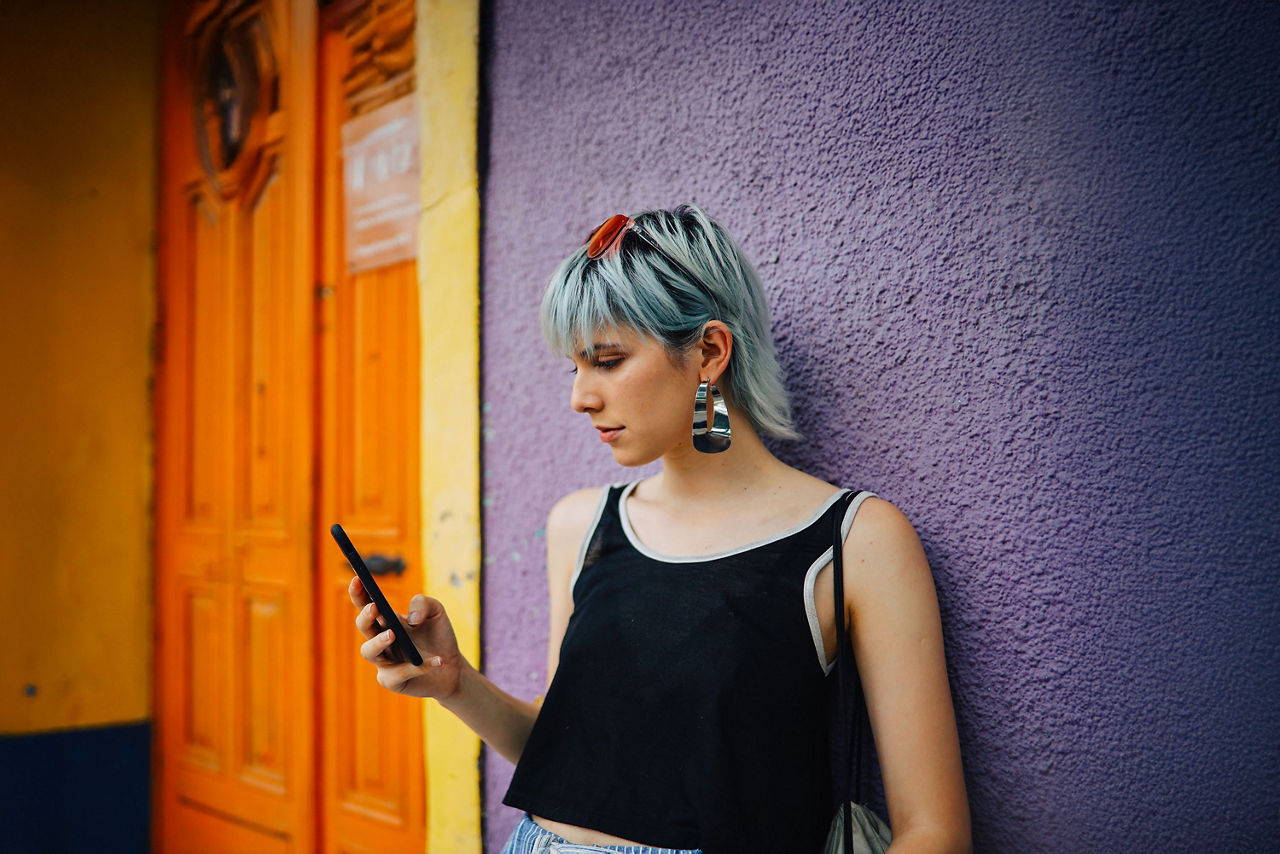 Junge Frau mit kurzen Haaren steht vor einer Hauswand und schaut auf ihr Handy