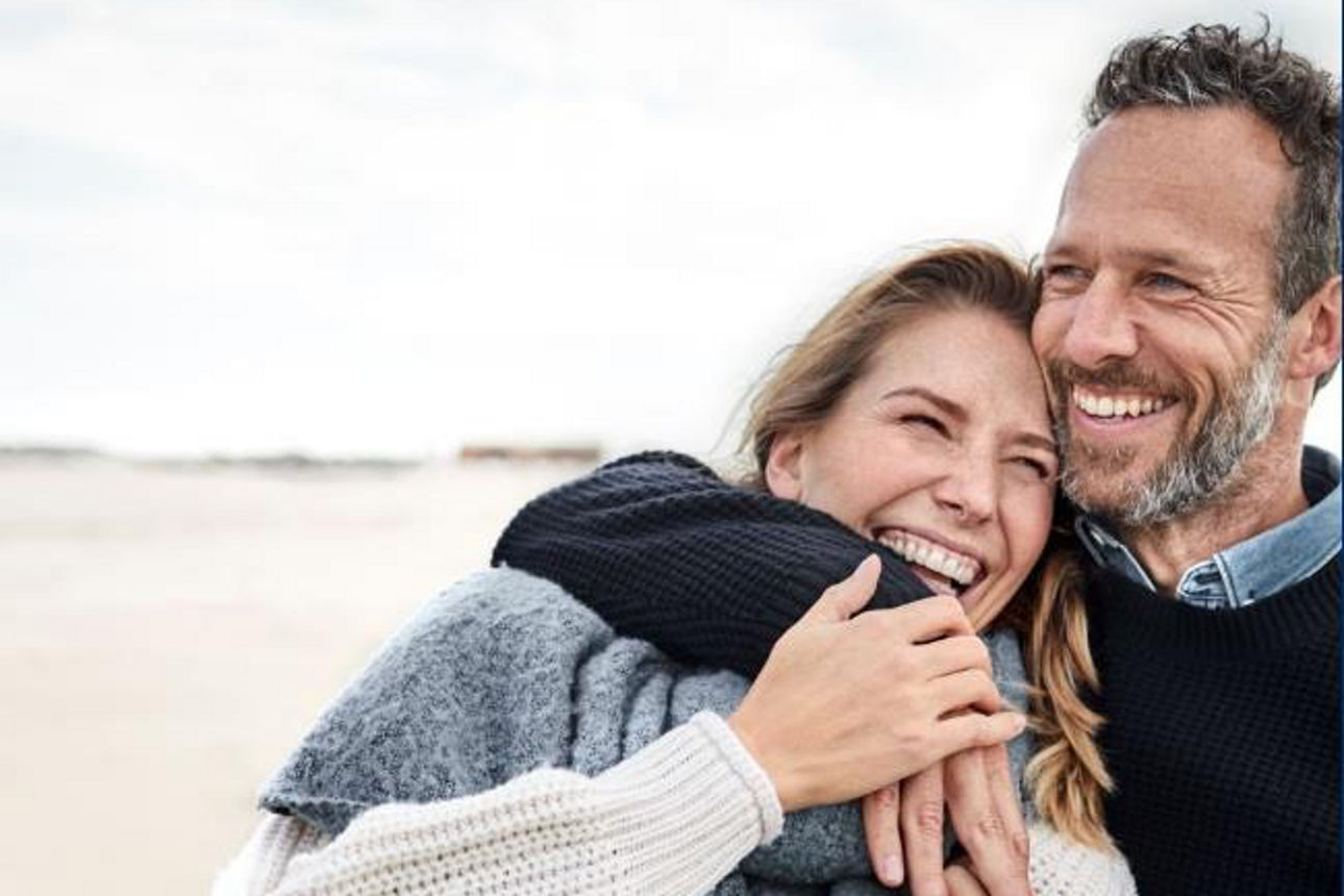 Eine Frau und ein Mann stehen am Strand, umarmen sich und lachen in die Blickrichtung des Bildbetrachters