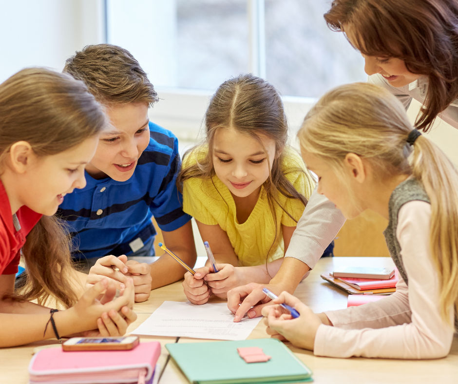 Vier Kinder und eine Lehrerin arbeiten gemeinsam an einem Tisch mit Heften und Stiften in einem Klassenraum