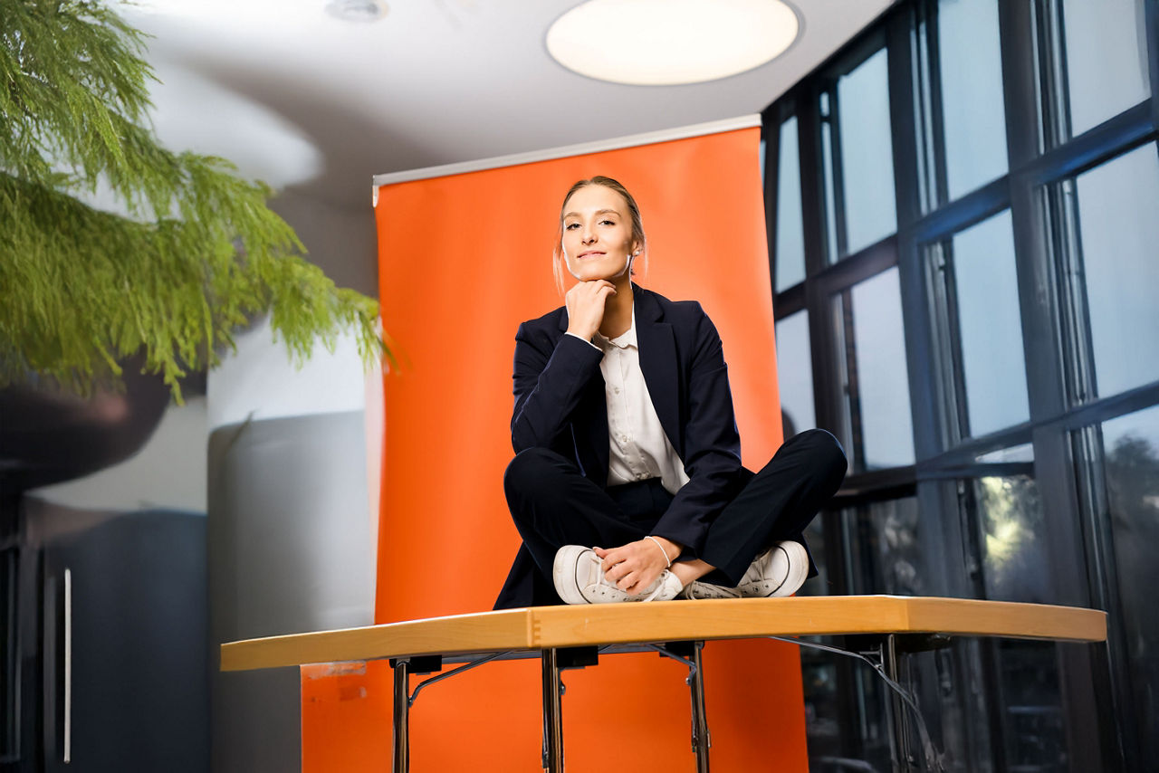 Die Auszubildende Lea Pfister sitzt vor einem orangefarbenem Rollup und stützt Kopf auf die Hand
