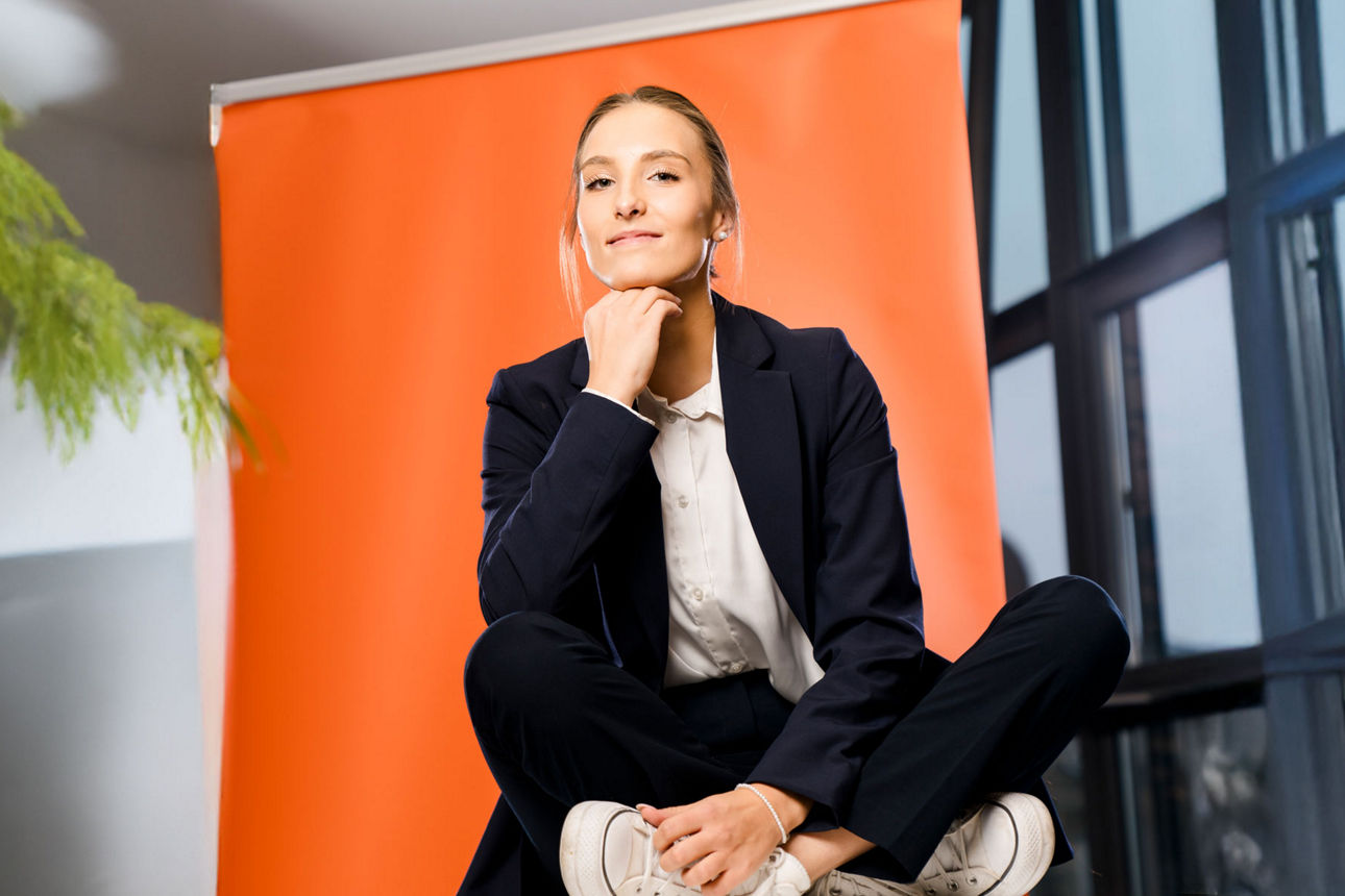 Die Auszubildende Lea Pfister sitzt vor einem orangefarbenem Rollup und stützt Kopf auf die Hand
