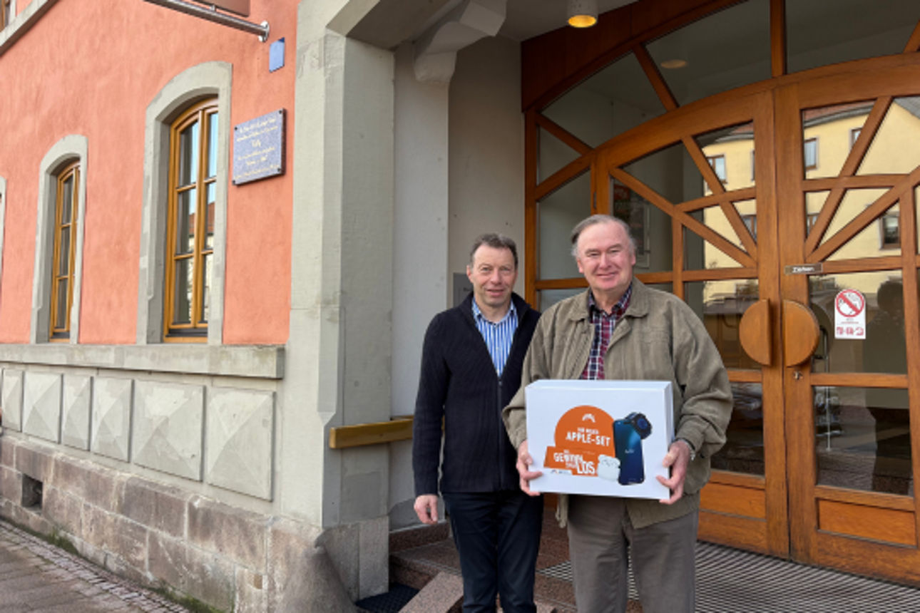 Zwei Männer stehen vor dem Eingang einer Bank. Der ältere Mann hält eine Box mit der Aufschrift "Apple-Set Gewinnsparlos.