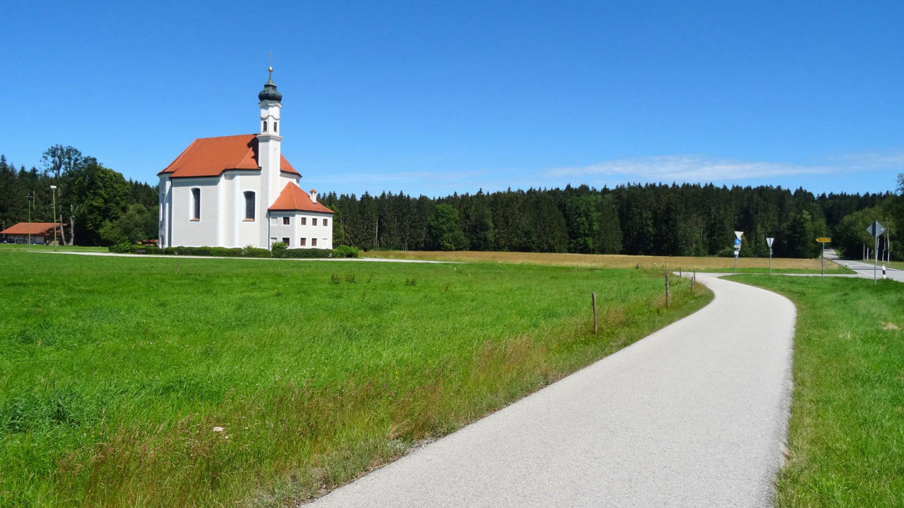 Eine weiße Kapelle mit einem roten Dach und einem Zwiebelturm steht inmitten einer weiten grünen Wiese. Ein geschwungener Weg führt von rechts unten zur Kapelle. Im Hintergrund erstreckt sich ein dunkler Wald unter einem strahlend blauen Himmel.