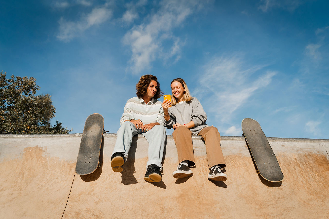 Skate colleagues sitting on top of a half pipe smiling and using phone while spending time together. Blue skay in the background.