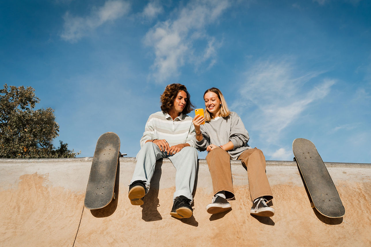 Skate colleagues sitting on top of a half pipe smiling and using phone while spending time together. Blue skay in the background.