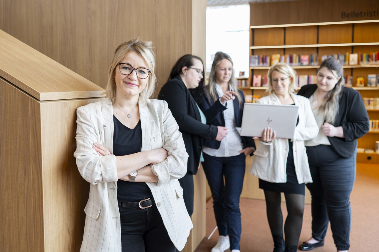 Das Team Digitale Beratung, bestehend aus 4 Frauen, steht mit Laptop beratend vor einem Bücherregal.