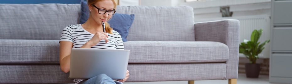 Frau mit Laptop sitzt vor einem Sofa und hält einen Stift in der Hand