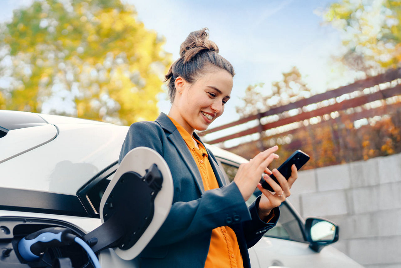 Frau vor einem Auto an der E-Tankstelle, die auf ihr Smartphone schaut