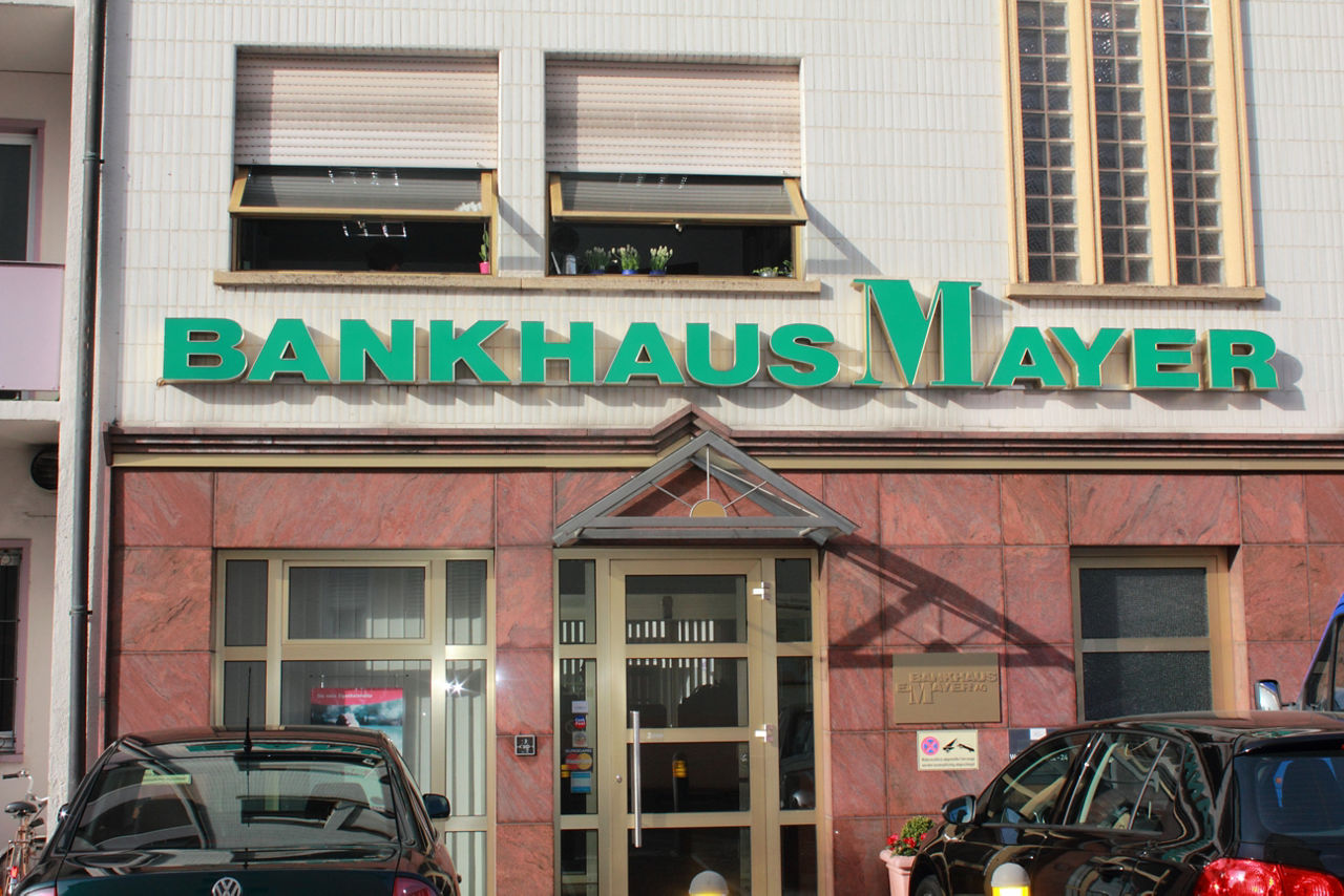 Auf dem Bild ist eine Filiale des Bankhaus Mayer von Vorne zu sehen. Über dem Eingang ist das Logo des Bankhauses in grün erkennbar. 
